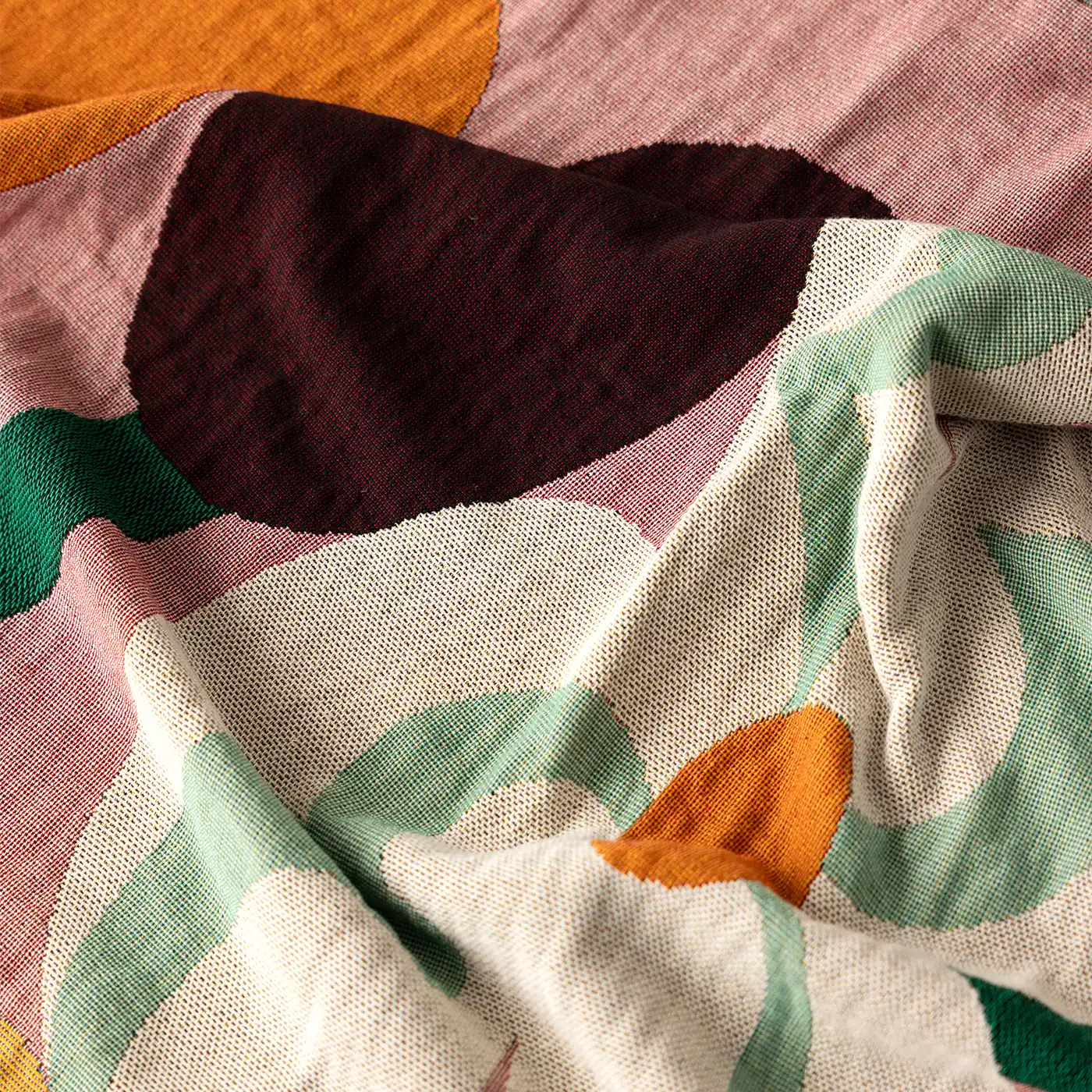 A close-up of a woven textile with a large-scale abstract floral or leaf pattern in muted pink, cream, sage green, orange, and deep burgundy.