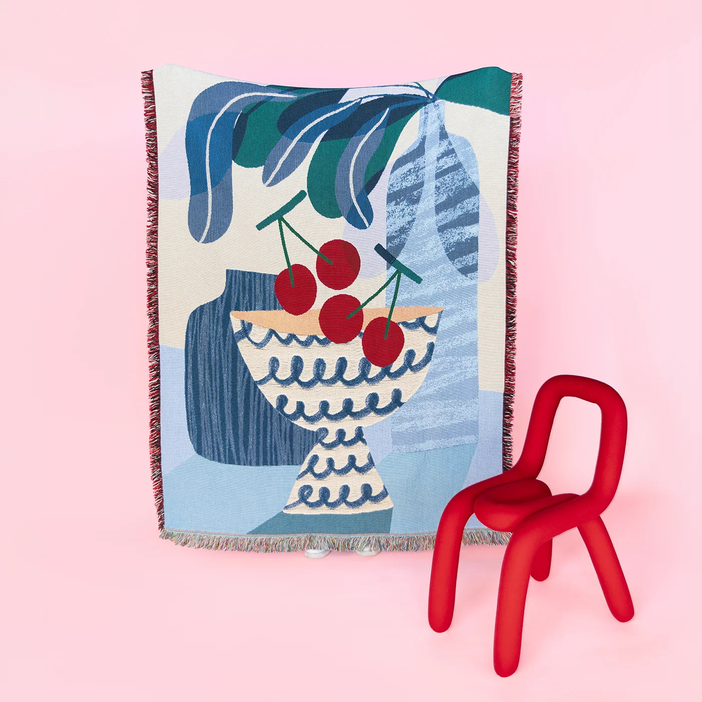 Decorative artist- designed throw blanket with a vase and cherry design on a pink background, accompanied by a red chair.