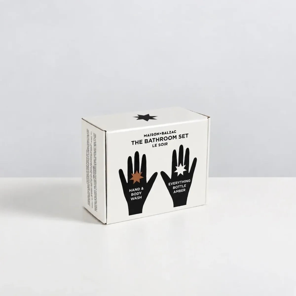 A white cardboard box for Maison & Balzac The Bathroom Set Le Soin, featuring black text and hand graphics on the front, photographed on a clean white surface against a light gray background.