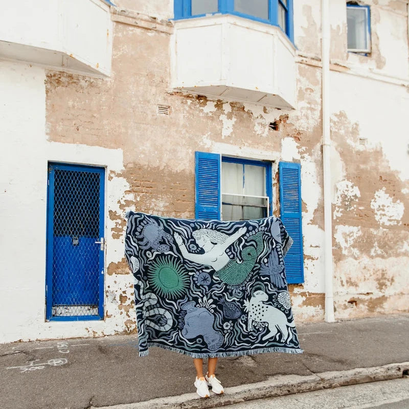 Person holding a large blue slowdown studio blanket with an Ancient Greek design in front of a building with blue shutters.