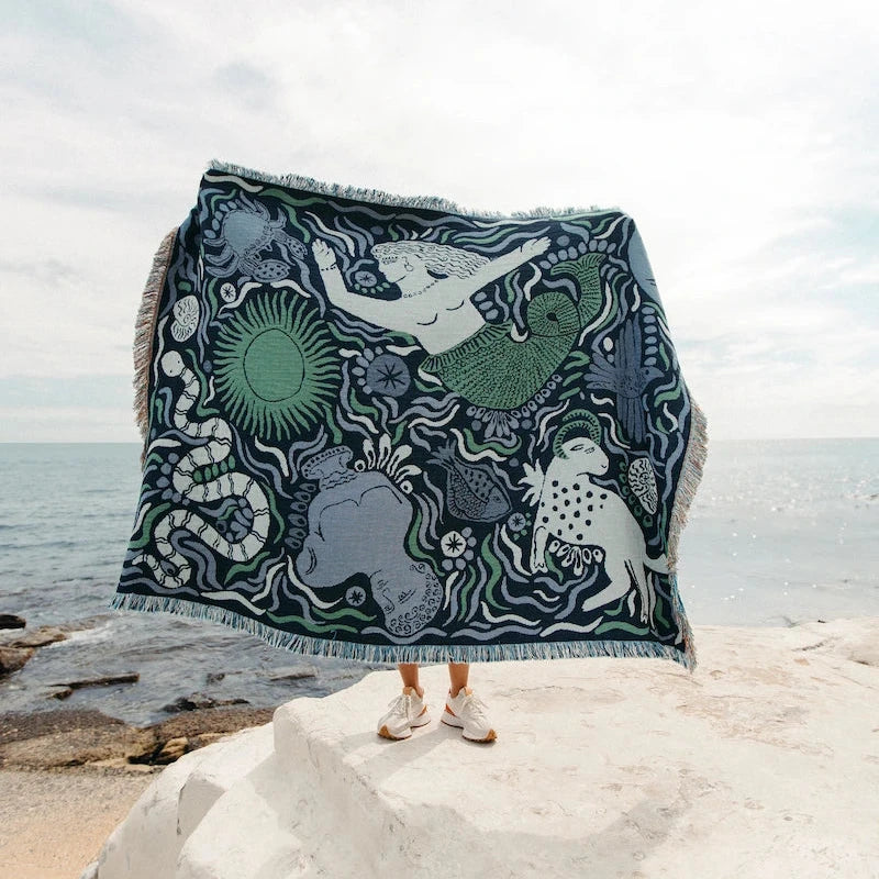 Person holding a decorative blue throw blanket with mermaid and sea creature pattern on a rocky beach.
