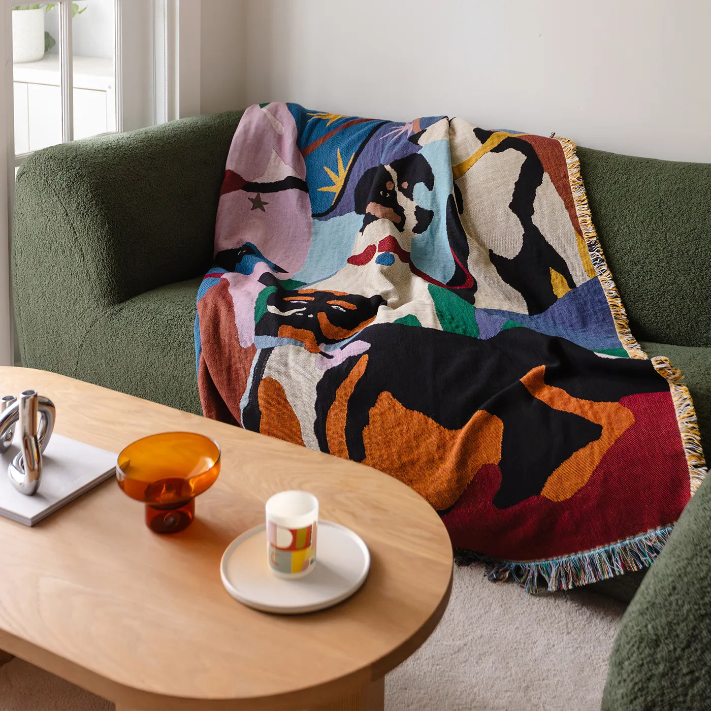 Colorful abstract-patterned throw blanket draped over a green sofa with a wooden coffee table in the foreground.