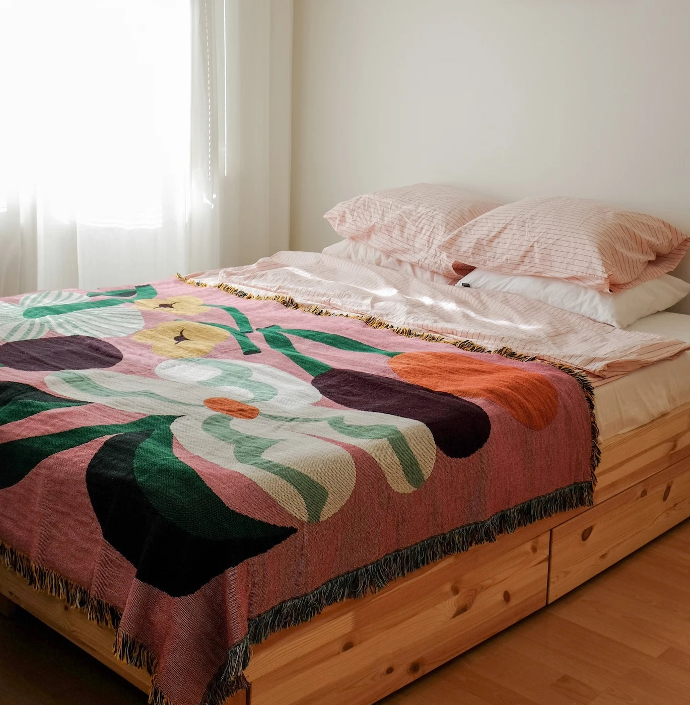 A pink floral woven blanket with green leaves and dark purple flower shapes is draped over a wooden bed frame in a softly lit bedroom. The bed is styled with light pink pillows and white bedding.
