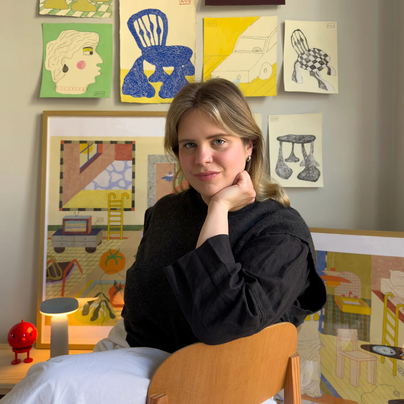 Polish artist Agata Malinowska sits in a wooden chair in a room, wearing a black long-sleeve top and white pants. Behind her, a wall is decorated with various colorful framed art prints and drawings.