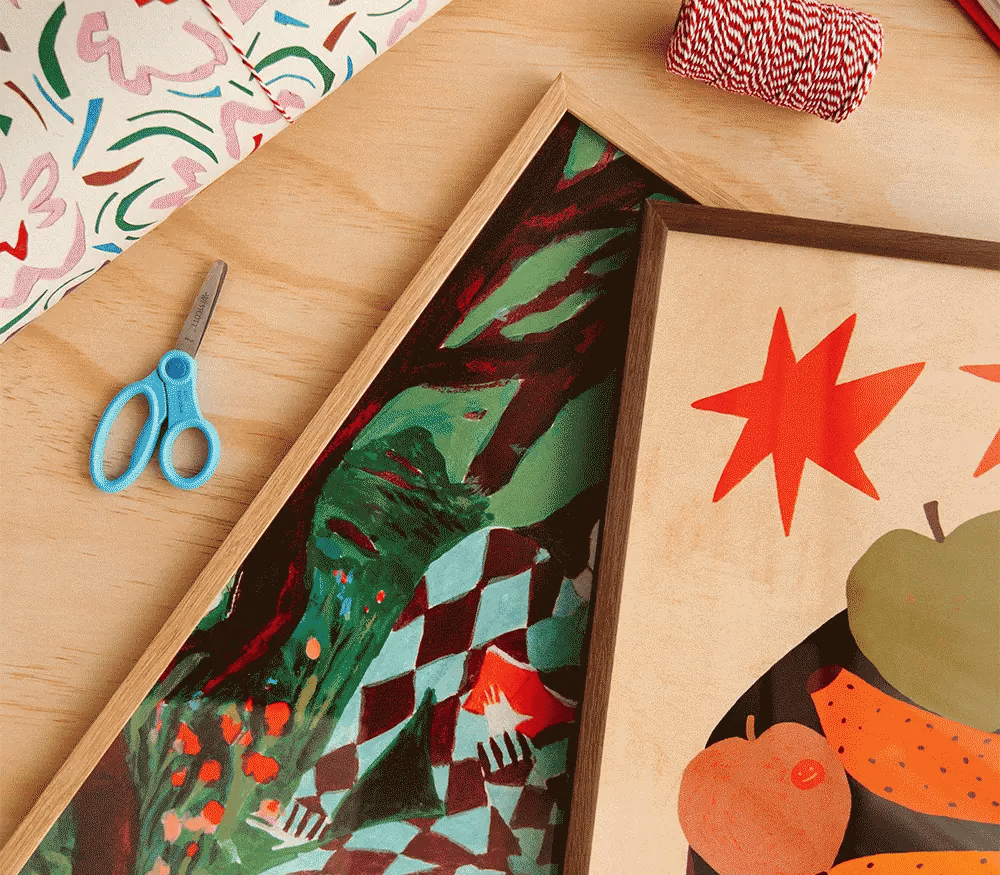 A styled flat-lay of framed art prints on a light wood surface, alongside blue-handled scissors, patterned paper, and red-and-white waffle twine. The visible artwork includes abstract and colorful illustrated scenes, with one print showing red star shapes and another showing green, red, black, and white geometric patterns.