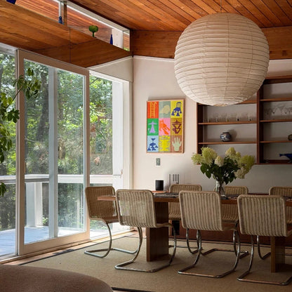 A modern dining room interior with a wooden dining table and six woven-back chairs, under a large white paper lantern pendant light. The room features a wood-paneled ceiling, a colorful geometric wall art print, built-in wooden shelves, and large glass sliding doors looking out onto greenery.
