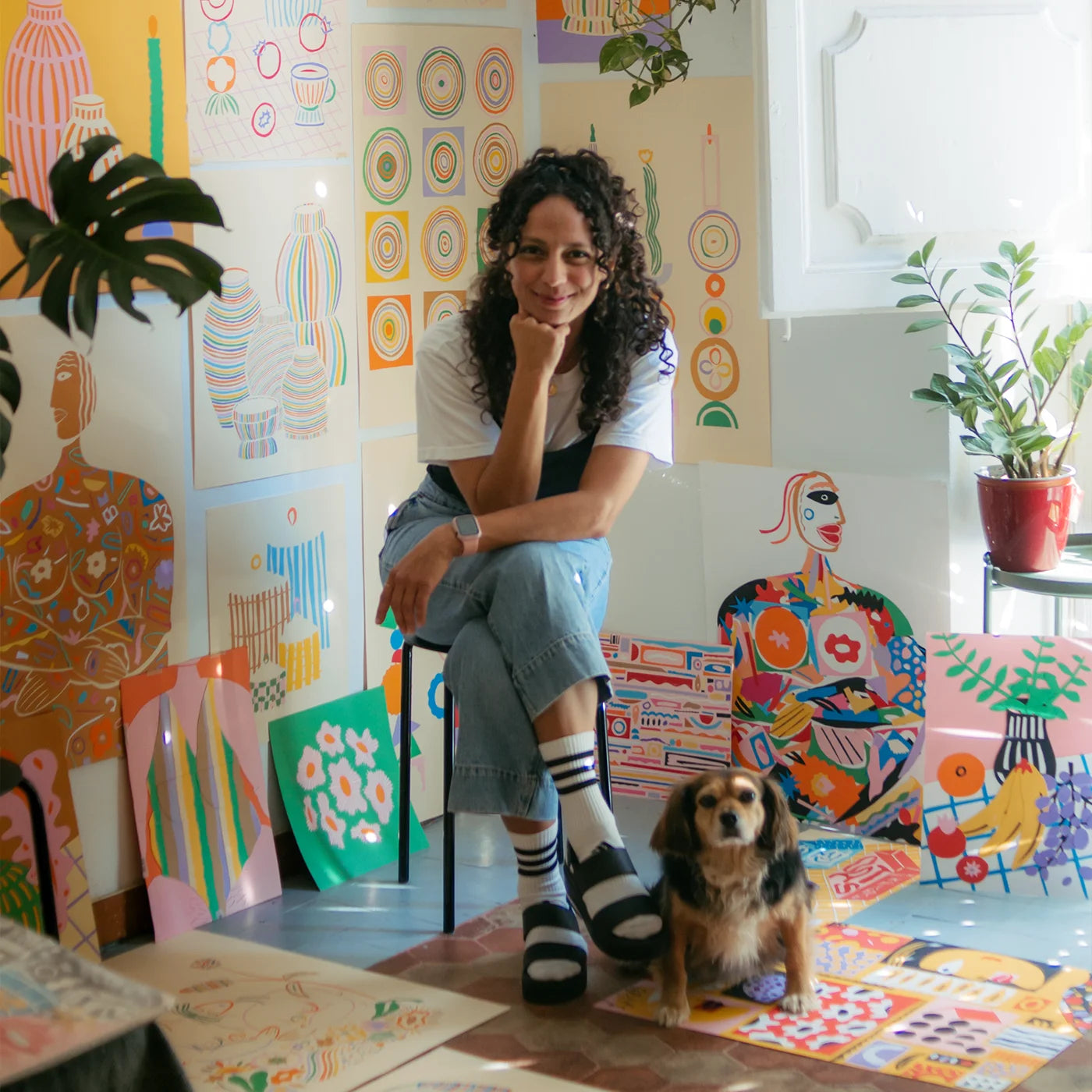 Spanish artist Carolina Restrepo sits on a stool in a brightly lit room surrounded by colorful art prints and paintings, with a small dog standing on a mat in front of her.