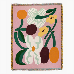 Slowdown Studio Cooper woven throw blanket with a pink background and a retro floral pattern in orange, yellow, white, and dark green. The blanket features fringed edges on all sides.
