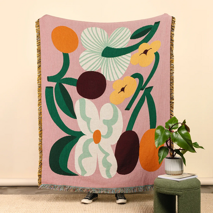 Slowdown Studio Cooper Throw. A pink woven throw blanket with a retro floral pattern is held up in a room setting. The blanket features large abstract flowers and leaves in green, white, yellow, orange, and dark brown, with fringed edges.