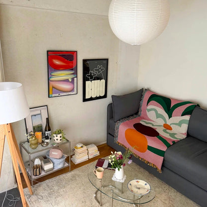 A cozy living room corner featuring a gray sofa with a colorful pink, green, and orange abstract floral throw blanket, a glass coffee table, a white paper lantern pendant light, and framed abstract wall art.
