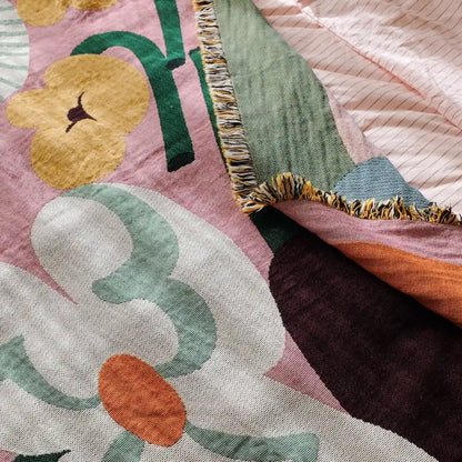 A close-up of a woven textile with a large floral pattern in dusty pink, cream, mint green, mustard yellow, and orange, with fringe trim along the edge.