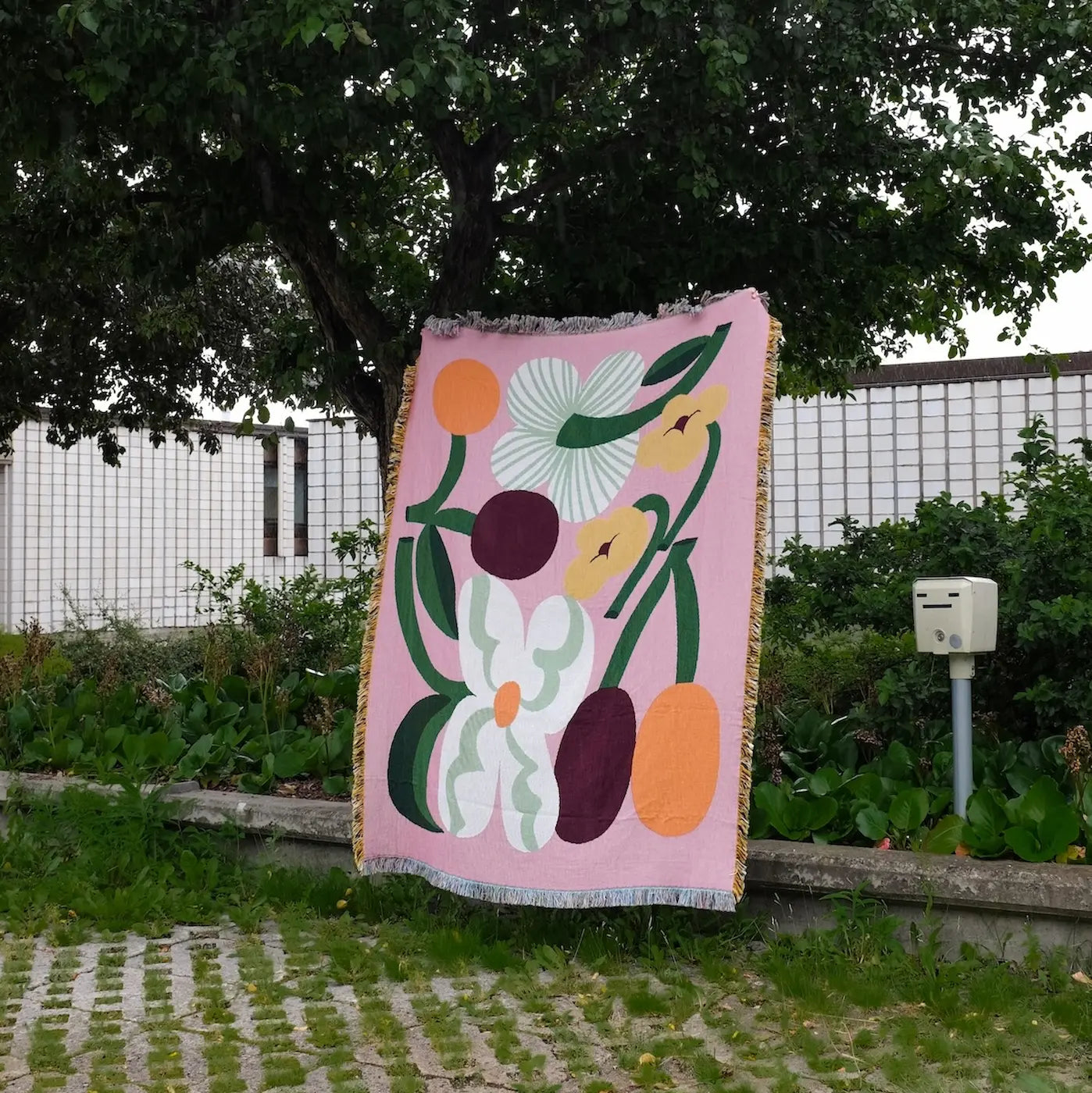A pink woven blanket with a retro floral pattern is draped outdoors against a tree in a garden setting.