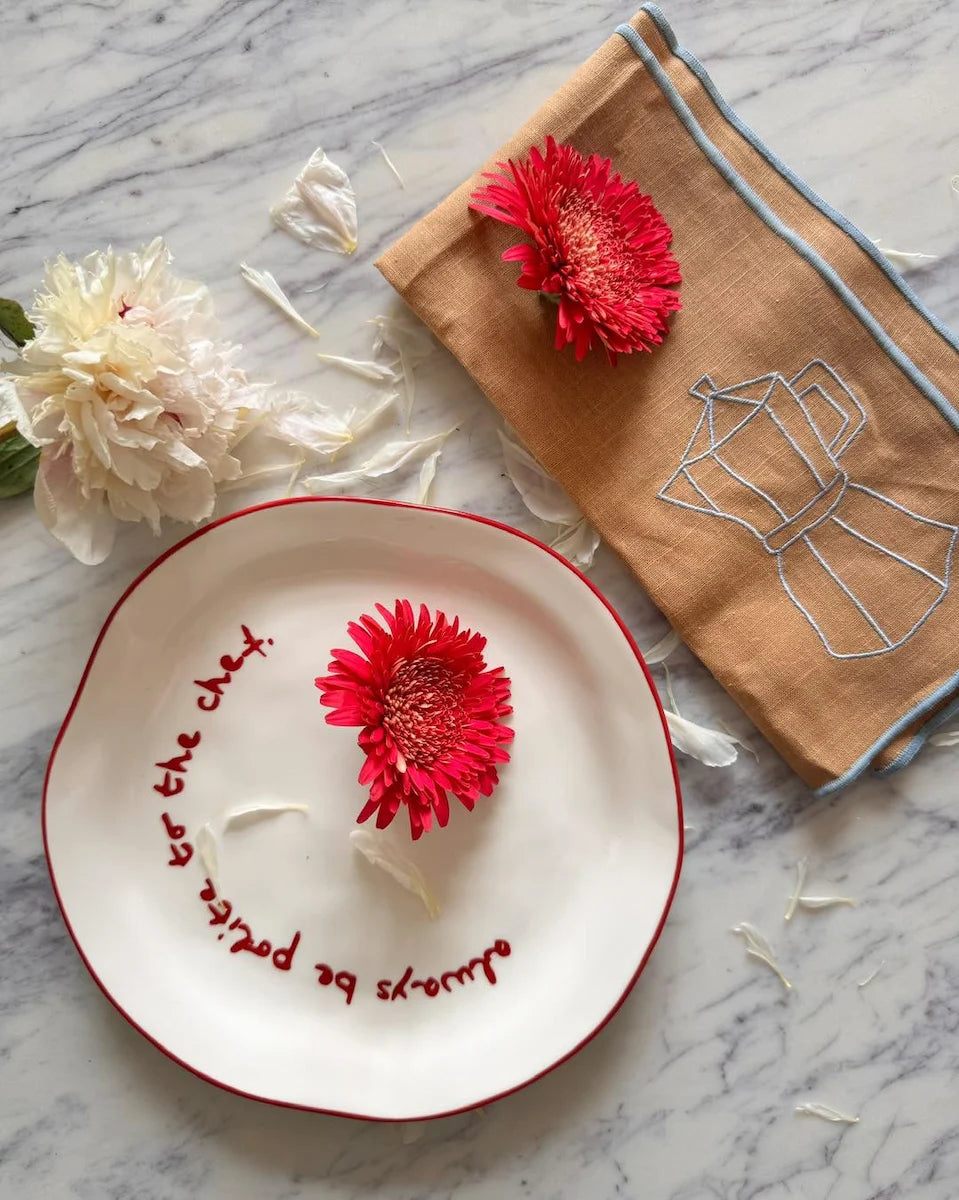 Decorative plate with a red flower and text, next to a brown pouch with a floral design on a marble surface.