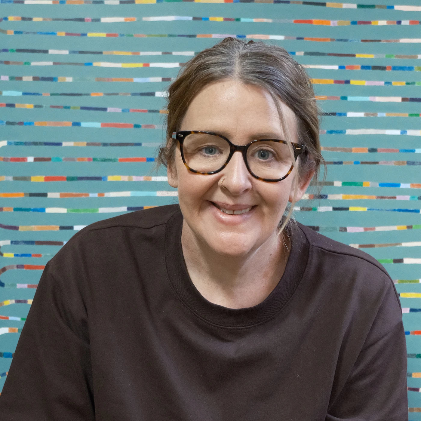 Australian artist Deb Chapman sits smiling with her pulled back, wearing black-rimmed glasses and a dark brown crewneck top, set against a colorful background of horizontal multicolored stripes.