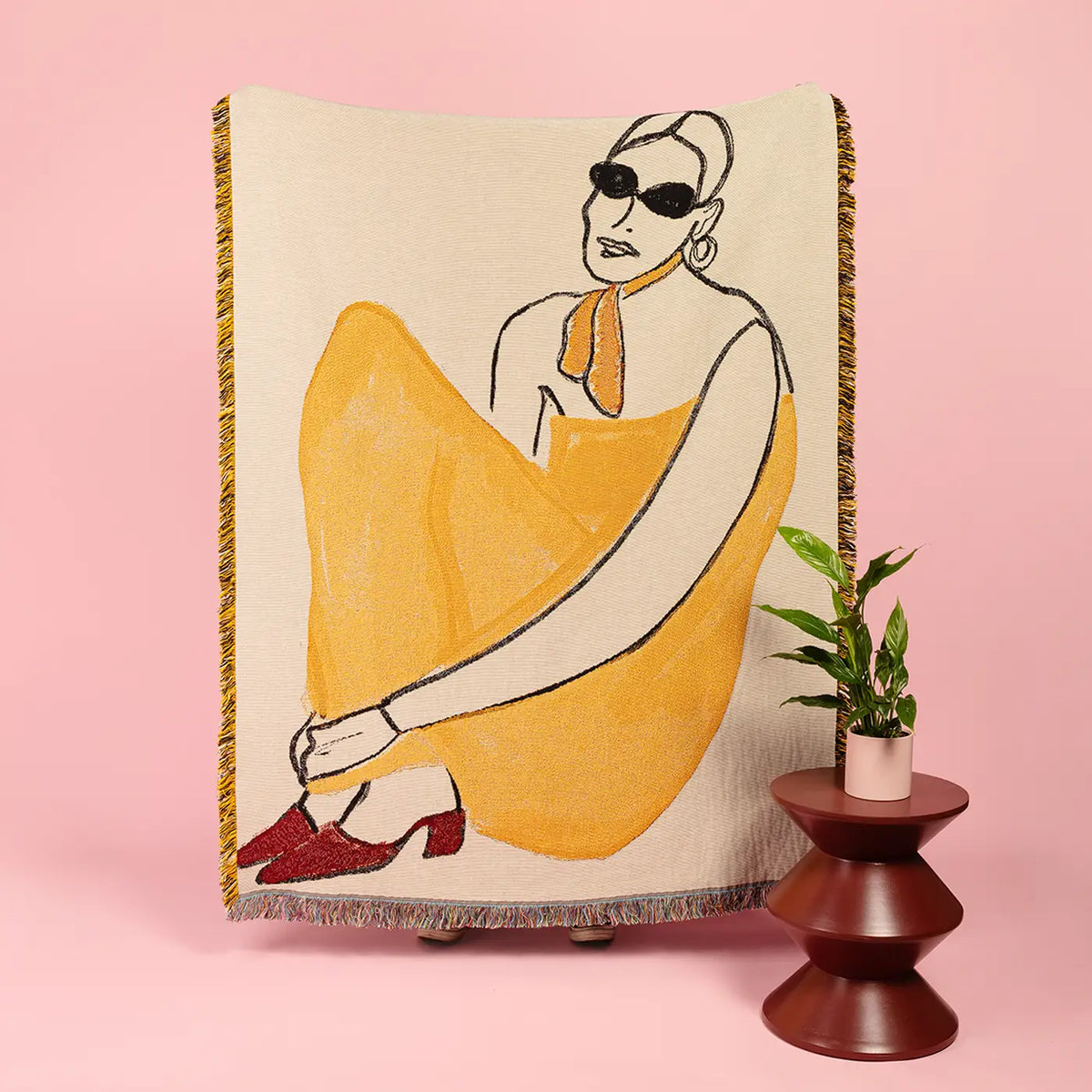 A woven throw blanket featuring a stylized illustration of a woman in a yellow dress and red shoes, displayed on a pink background next to a small potted plant on a wooden stand.