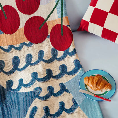 Slowdown Studio decorative blawoven throw with cherry pattern, checkered pillow, and plate of croissant on a couch.