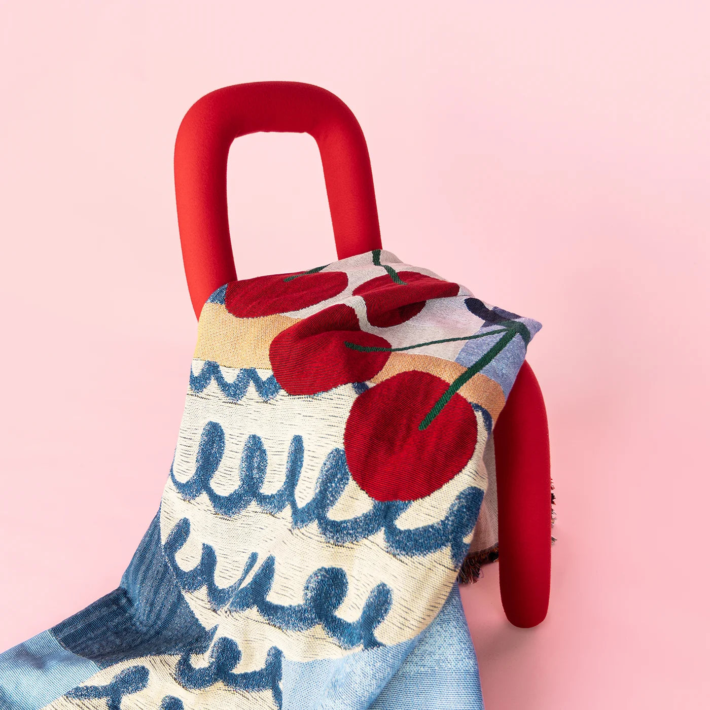 Artist-designed woven throw blanket with a blue vase &amp; cherry design, draped over a red chair on a pink background.