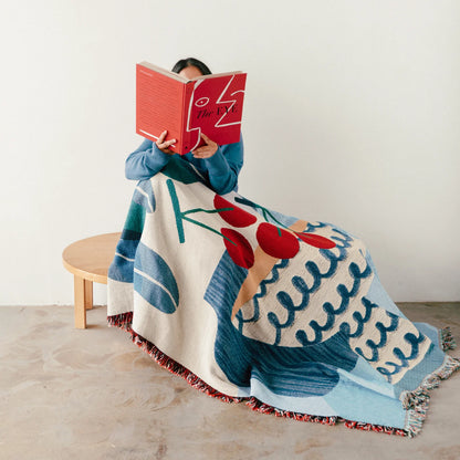 Person sitting on a chair with a colorful patterned blanket, reading a red book.