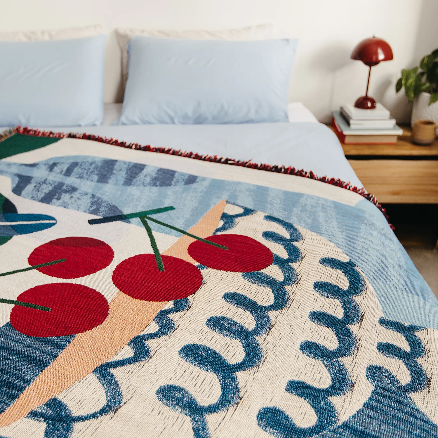 Artist-designed woven blanket with illustrative blue fruit bowl, abstract patterns and red cherries on a bed. 