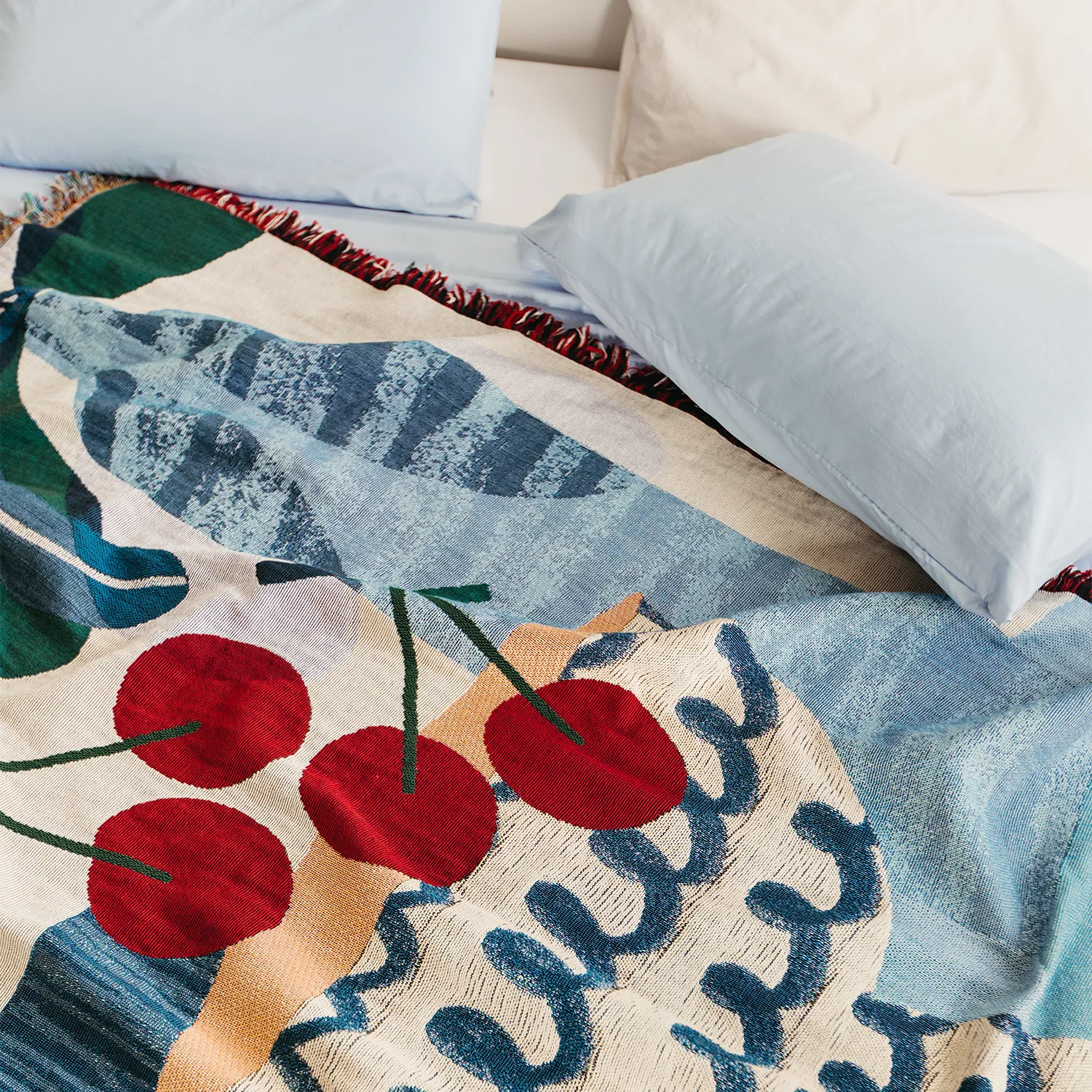 Slowdown Studio Freya throw on a bed. The blanket features an abstract blue illustration artwork with a fruit bowl containing red cherries and botanicals framing the composition. 