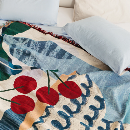 Slowdown Studio Freya throw on a bed. The blanket features an abstract blue illustration artwork with a fruit bowl containing red cherries and botanicals framing the composition. 
