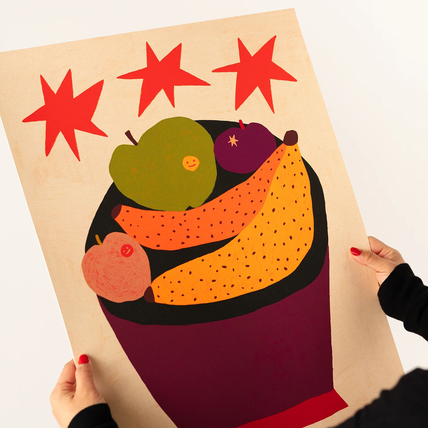 A person holds a large art print featuring a colorful fruit bowl illustration with red stars above. The print is displayed against a plain white background, with the artist's hands visible holding the corners of the paper.