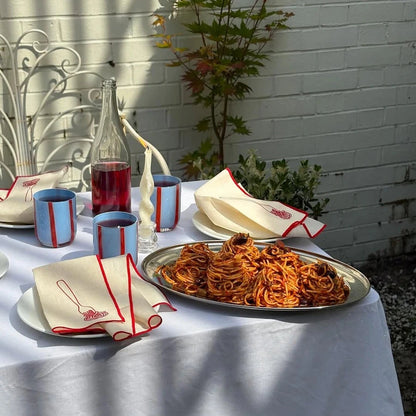 Outdoor table setting with spaghetti, napkins, cups, and a bottle against a white brick wall.