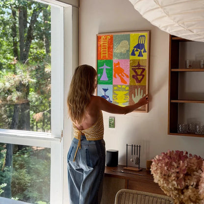 A woman in a beige top and blue jeans hangs a colorful framed art print on a beige wall in a modern room with a large window showing trees outside.