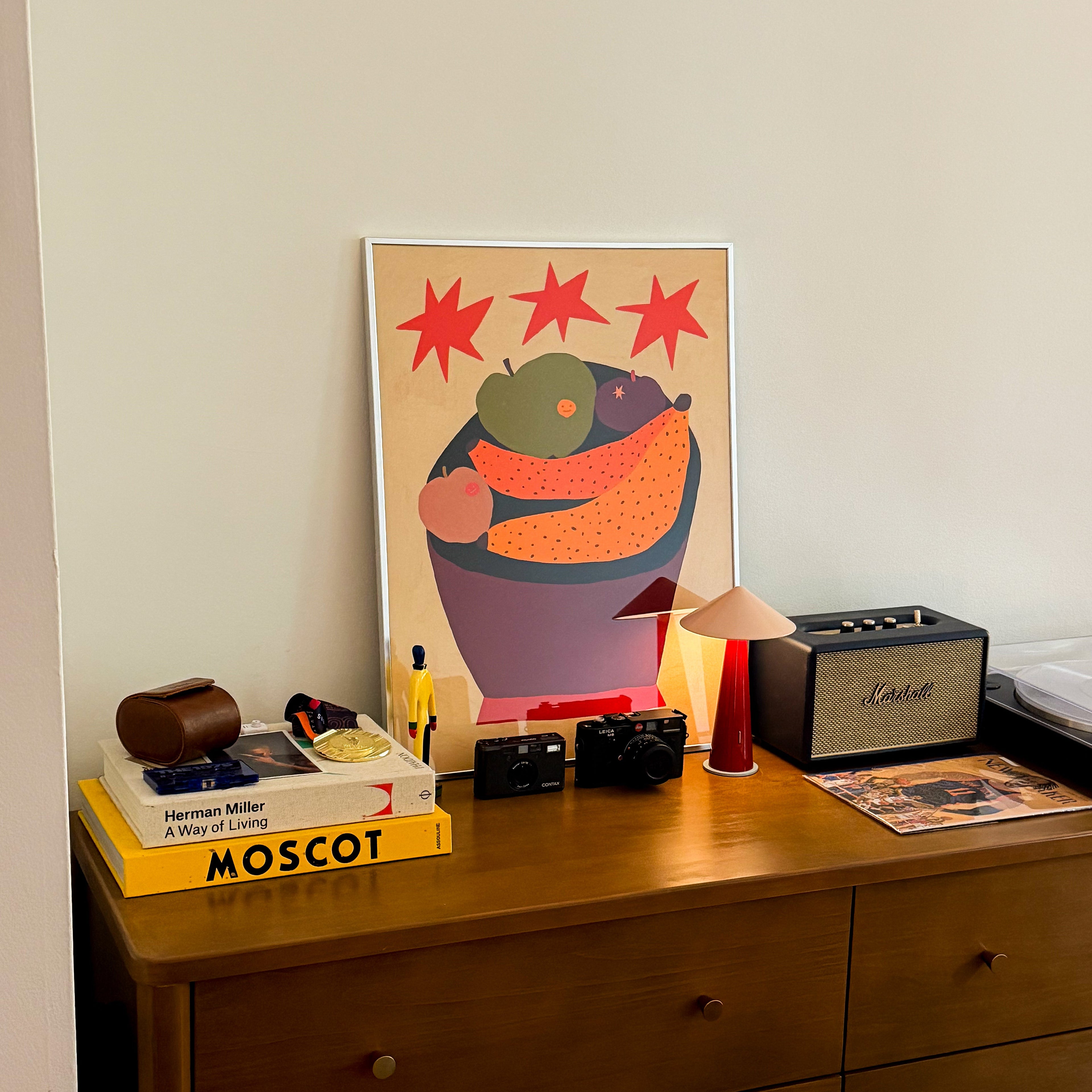 A framed art print featuring a colorful abstract illustration of a bowl with red stars, styled on a wooden dresser alongside a Marshall speaker, a small lamp, and a yellow book.