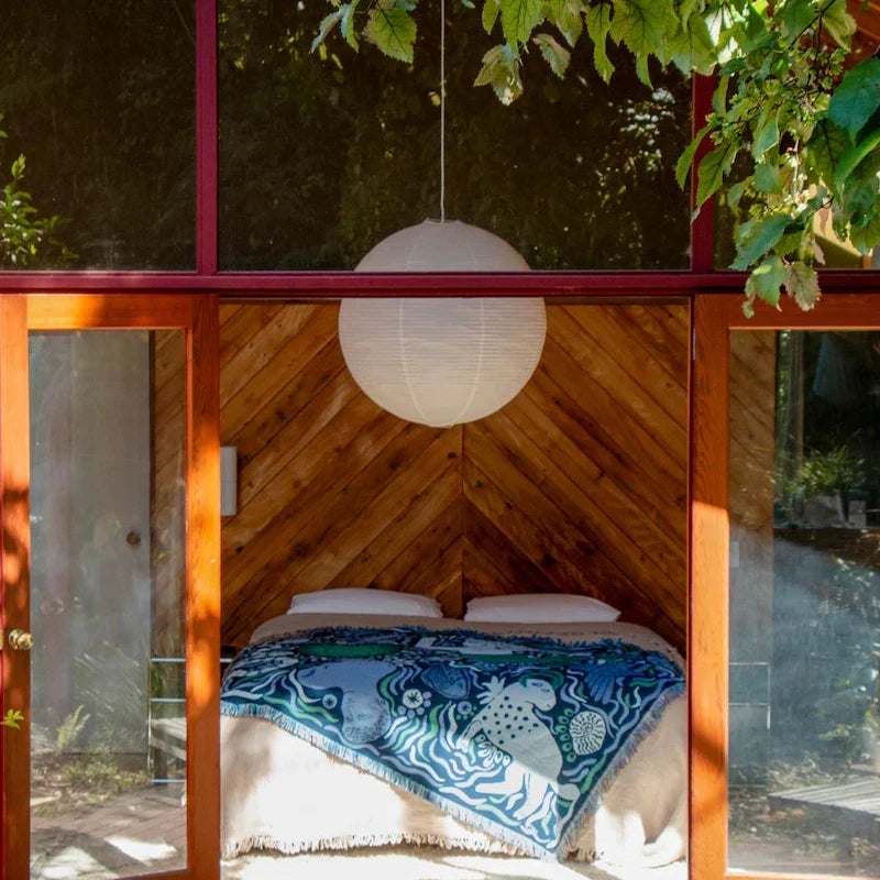 Wooden cabin with glass walls, bed, and hanging light fixture surrounded by greenery. A blue slowdown studio bed blanket is styled on the bed. 