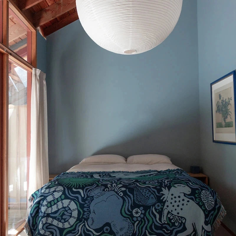 Bedroom with blue walls, blue slowdown studio bed blanket, wooden ceiling, and a large white pendant light.