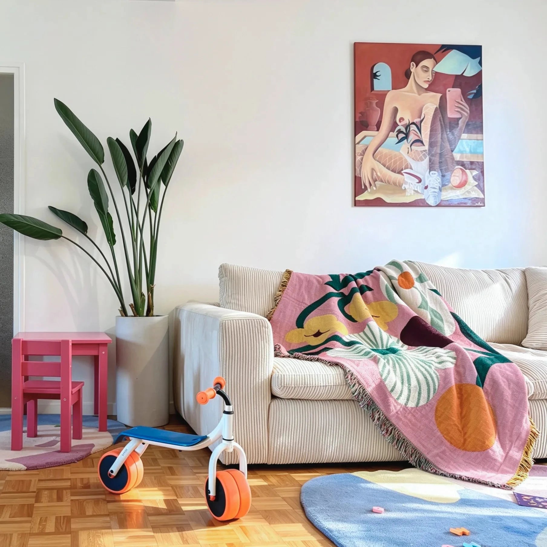 A styled living room scene featuring a beige corduroy sofa with a pink tropical leaf-patterned throw blanket, a small pink side table, a white tricycle with orange wheels, and a colorful framed painting on the wall.