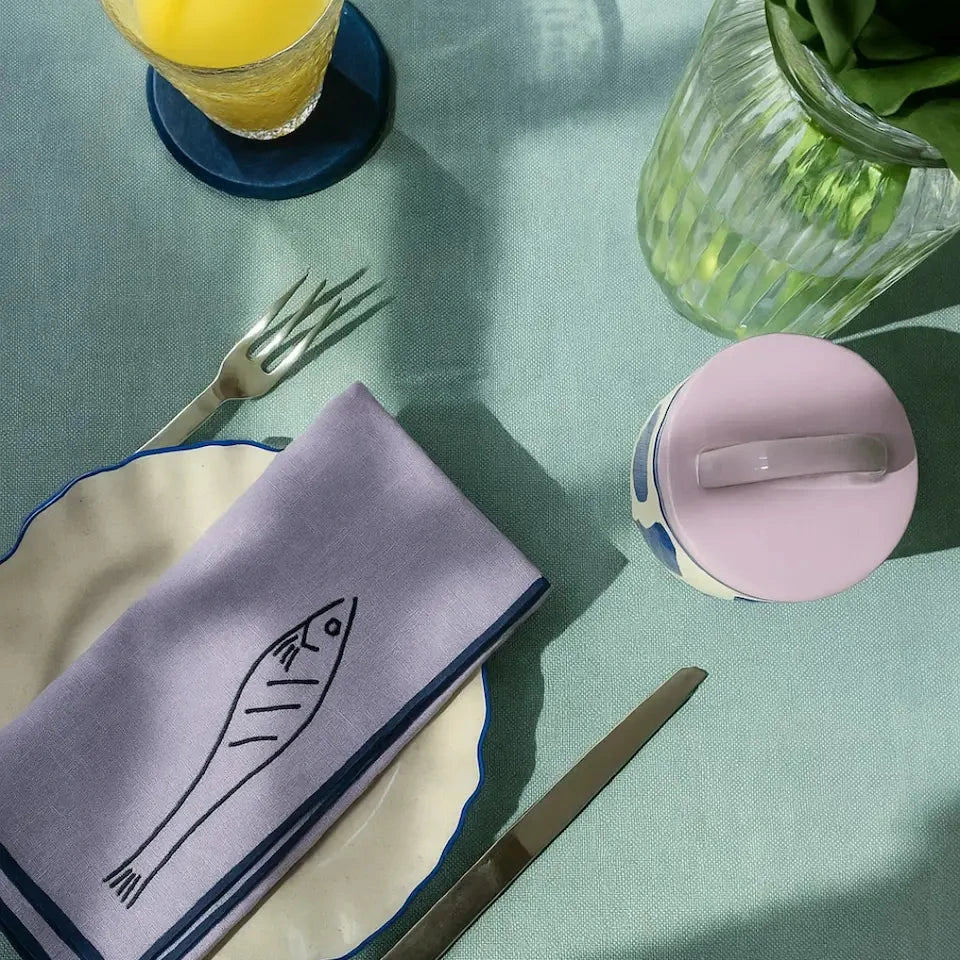 Table setting with a fish-themed napkin, glass of orange juice, and pink container.