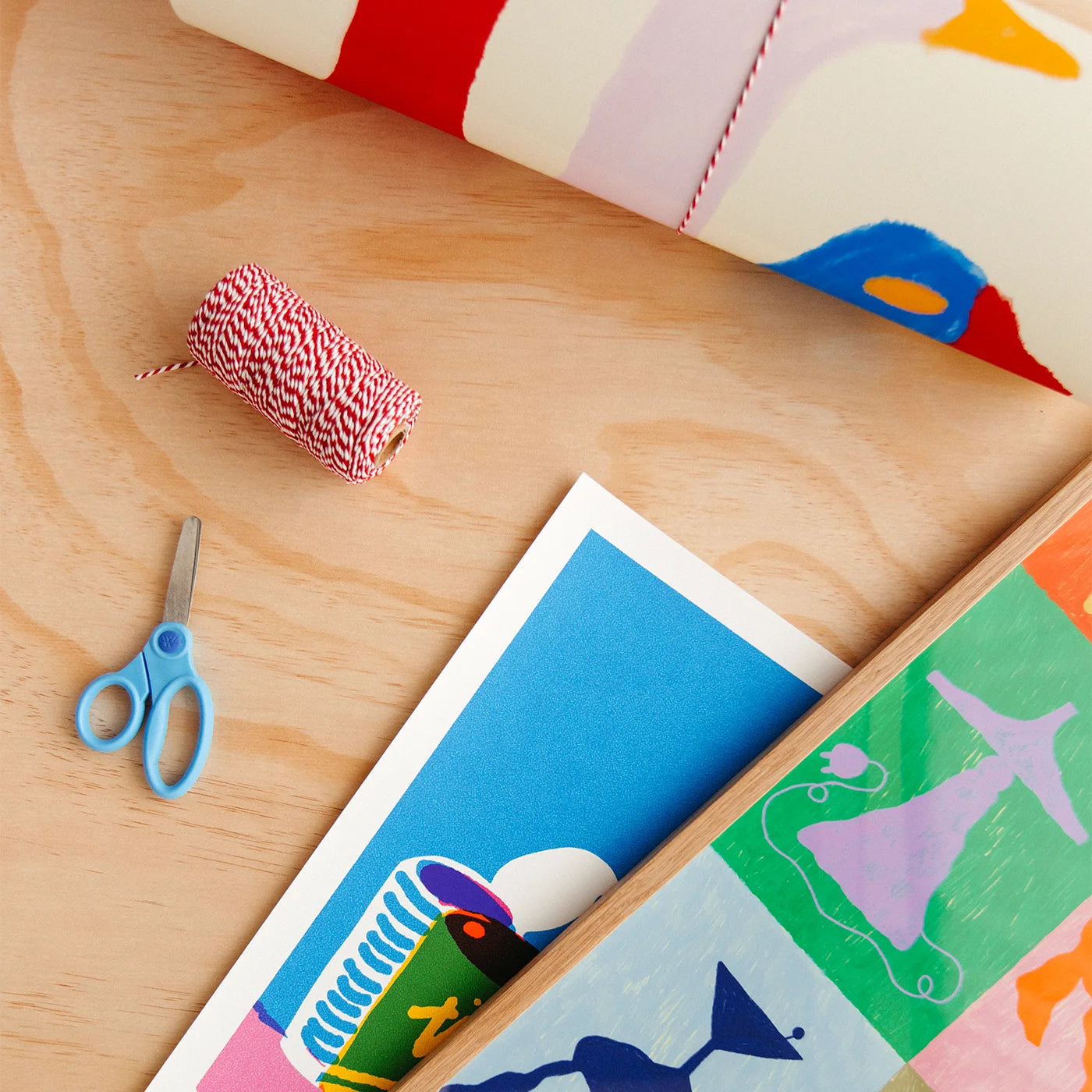 A styled flat-lay of colorful craft supplies on a light wood surface, including a spool of red-and-white bakers twine, blue-handled scissors, an art print, and a rolled sheet of patterned paper with red, white, pink, blue, and yellow sections.