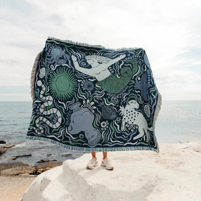 Person holding a decorative blue throw blanket with mermaid and sea creature pattern on a rocky beach.