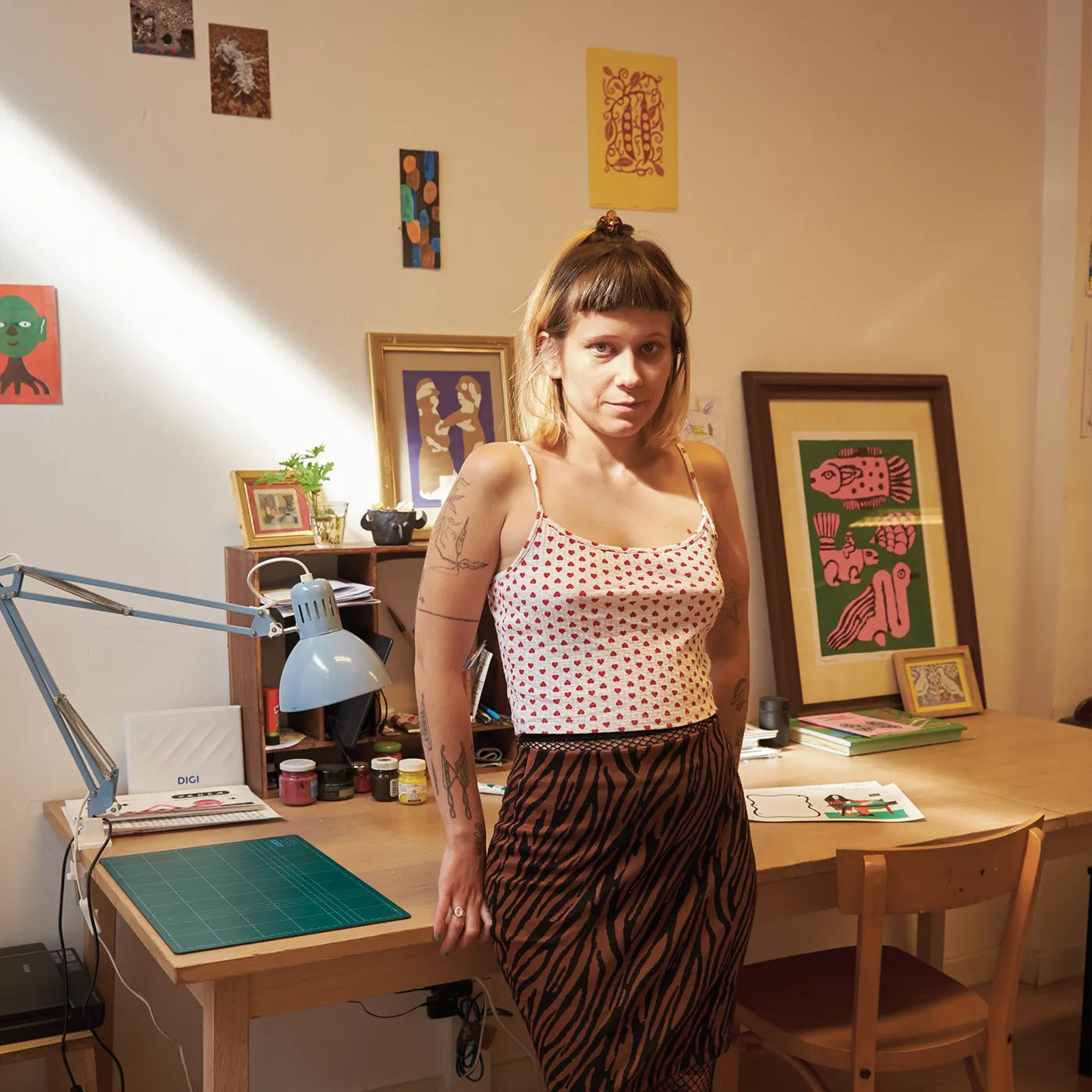 Spanish artist Nina Izycka stands in a sunlit art studio wearing a white camisole with red polka dots and a brown zebra-print skirt. She is positioned next to a wooden desk with a green cutting mat, a blue desk lamp, and framed colorful art prints on the beige walls behind her.