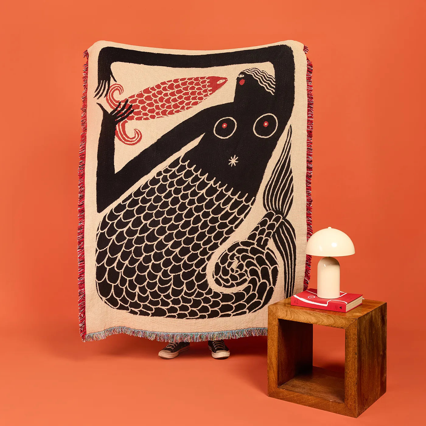 Decorative bed blanket in colors black, beige and red with a fish and mermaid figure design on an orange background. Folk art style.