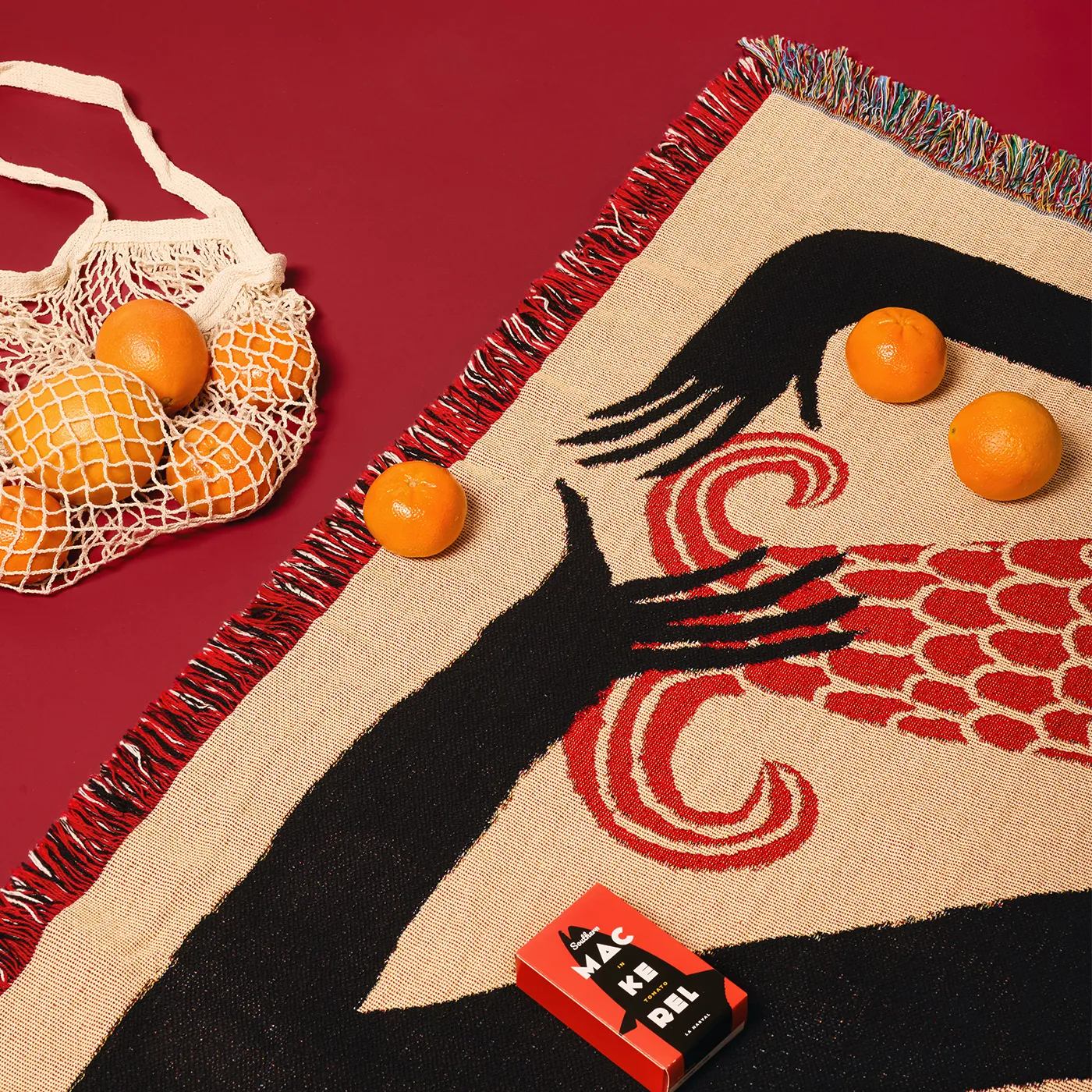 Woven blanket textile with artistic design of a hand reaching out, surrounded by oranges and a mesh bag on a red background.