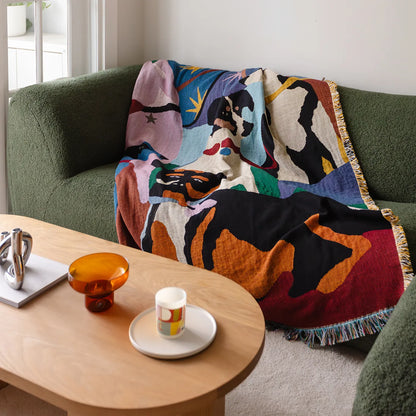 Colorful abstract-patterned throw blanket draped over a green sofa with a wooden coffee table in the foreground.