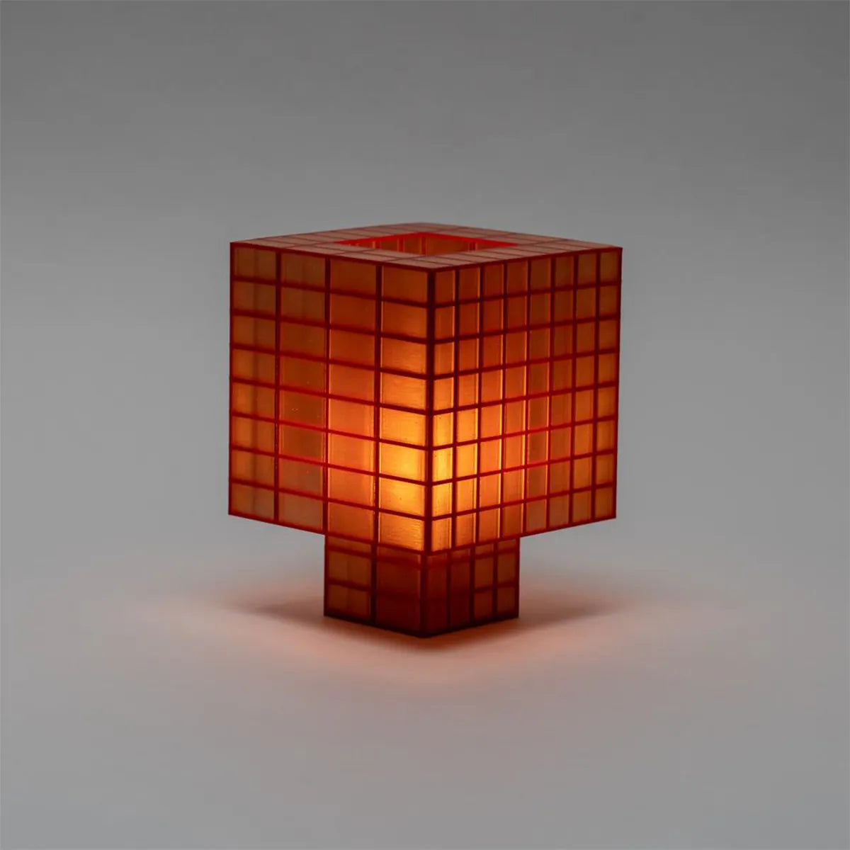 A Ridezign Tesser 8:3 table lamp with a translucent orange-red glass body and a glowing warm light inside, set against a neutral gray background.