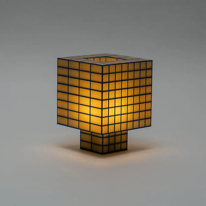 A Ridezign Tesser 8:3 table lamp with a cube-shaped shade and matching base, featuring a black grid pattern over warm yellow-gold translucent panels, set against a neutral gray background.