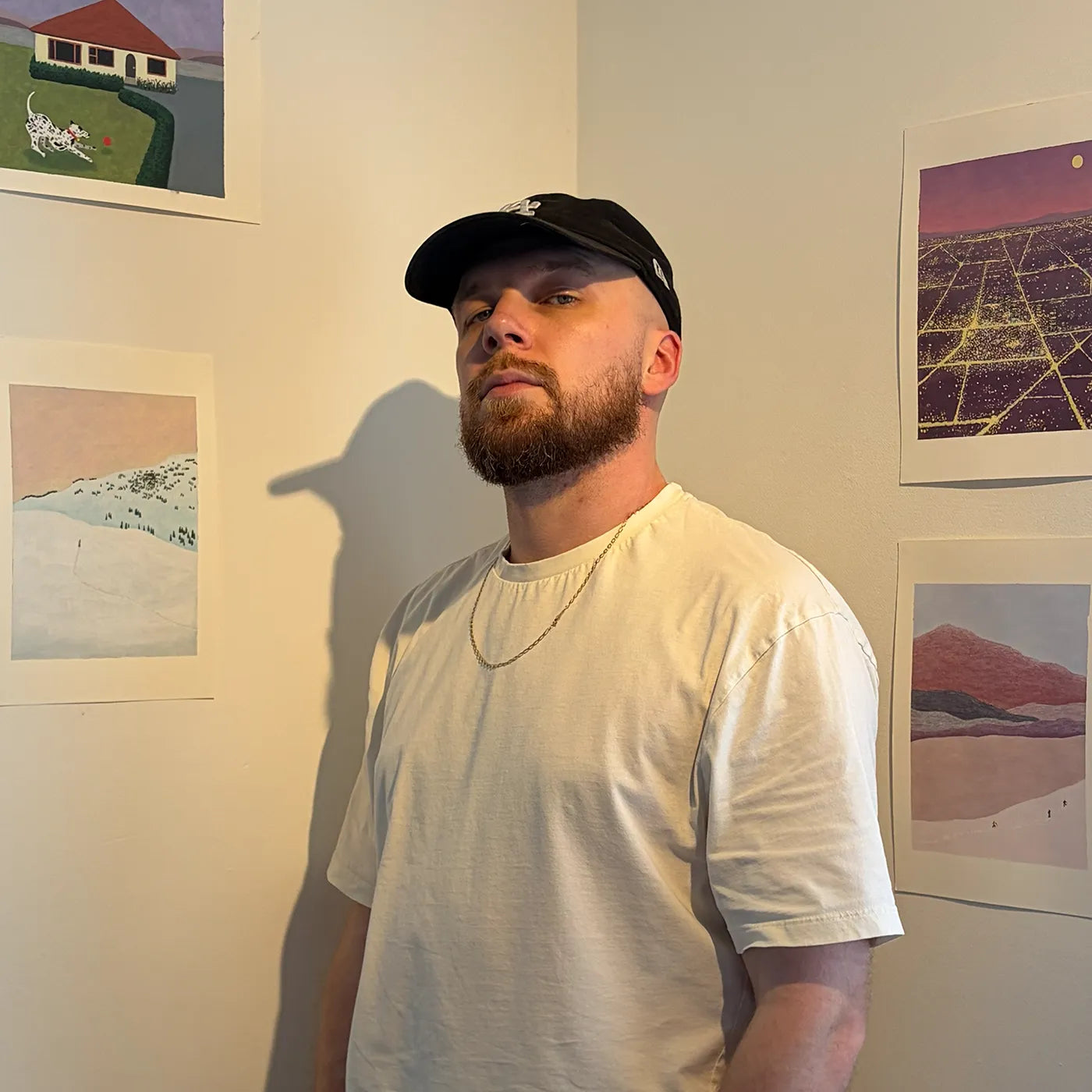 Portrait of English artist Ryan Bradley wearing a black baseball cap and a white t-shirt, standing in an indoor setting with framed art on the wall. The image is presented in a decorative scalloped circular frame with the text 'Ryan Bradley' underneath on a white background.