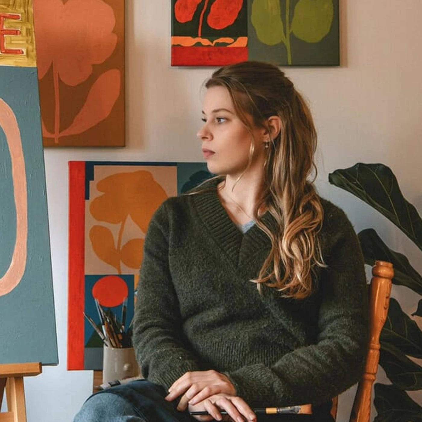 Portrait of English artist Sophie Deller sitting in a wooden chair wearing a dark olive green knit sweater, set against a cozy room decorated with colorful floral paintings and a potted plant.