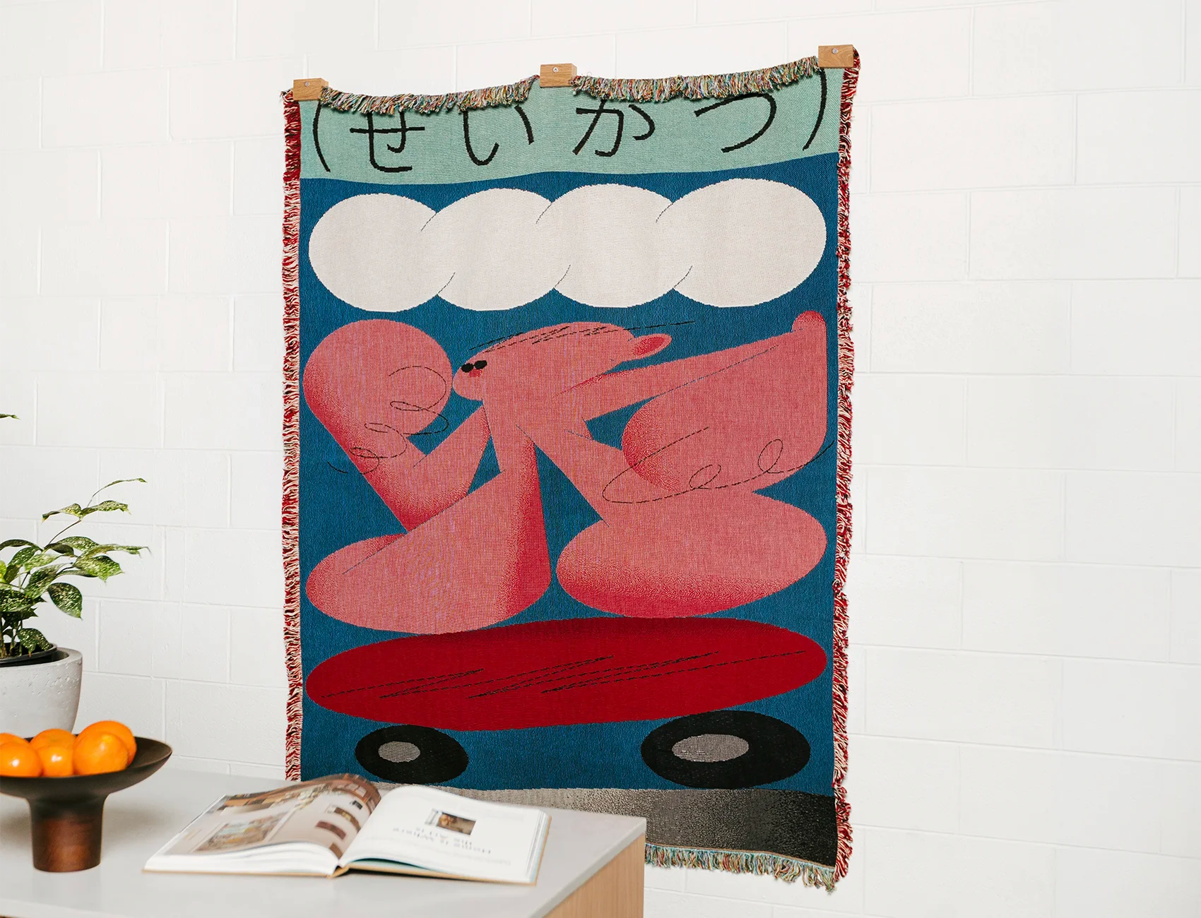 Woven tapestry with abstract design of a pink character on a blue background, displayed on a white wall, hung up with Slowdown Studio Blanket Hanger Clips.