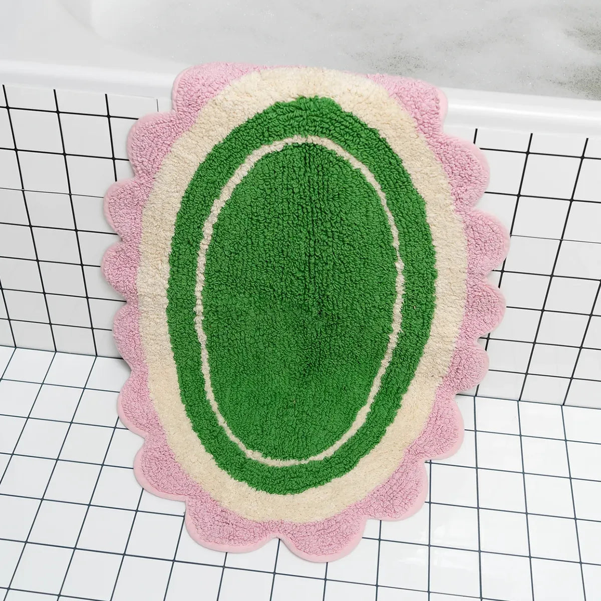 Decorative bath mat with green center, pink border, and scalloped edge on a grid background.