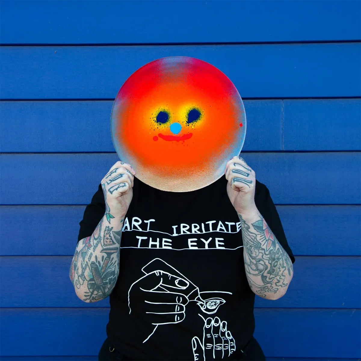 Person holding a colorful smiley face plate in front of their face against a blue wooden panel background.