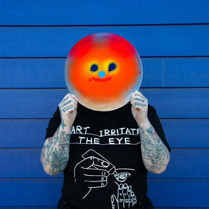 Person holding a colorful smiley face plate in front of their face against a blue wooden panel background.