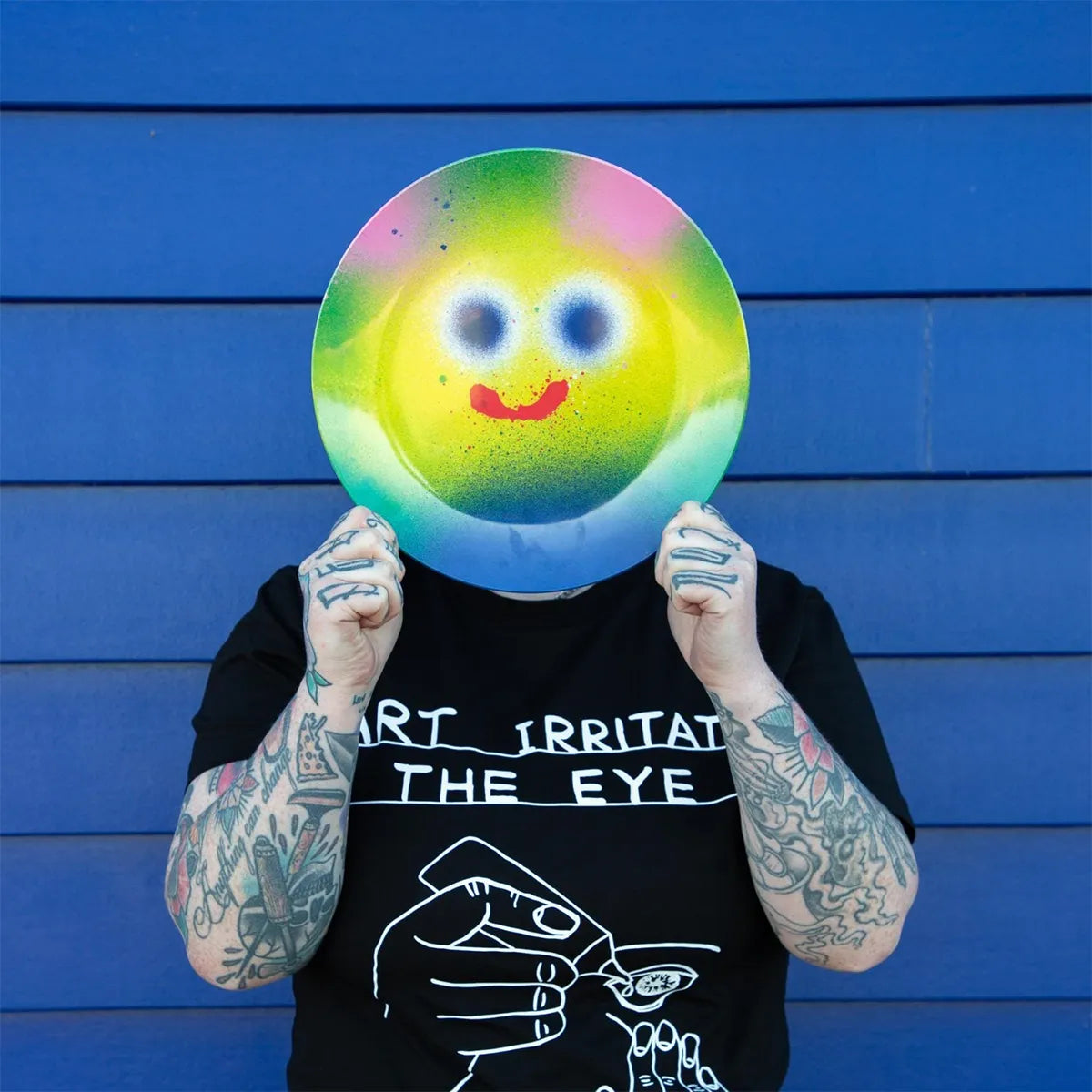Person holding a colorful smiley face plate in front of their face against a blue background