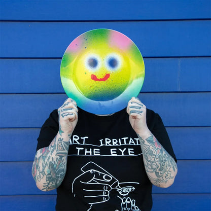 Person holding a colorful smiley face plate in front of their face against a blue background