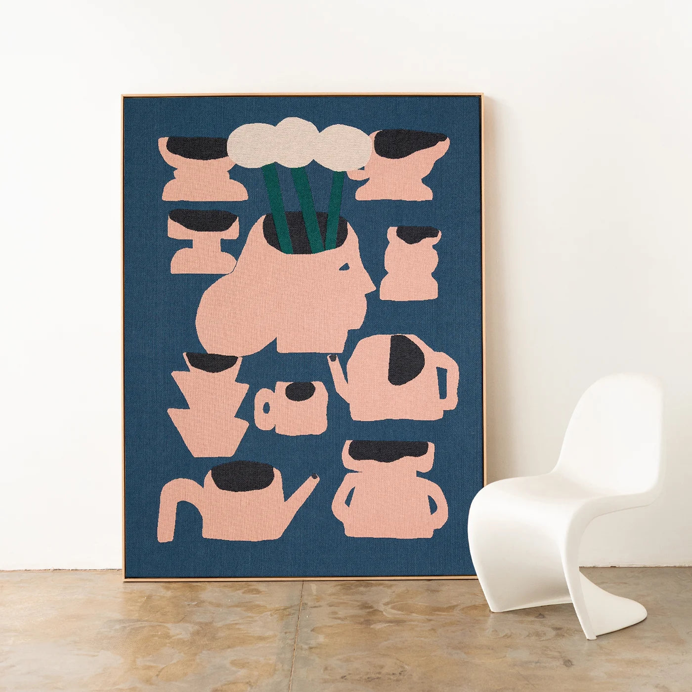 Framed abstract woven artwork with pink vase shapes on a blue background, displayed in a room with a white chair. Woven throw by Slowdown Studio.